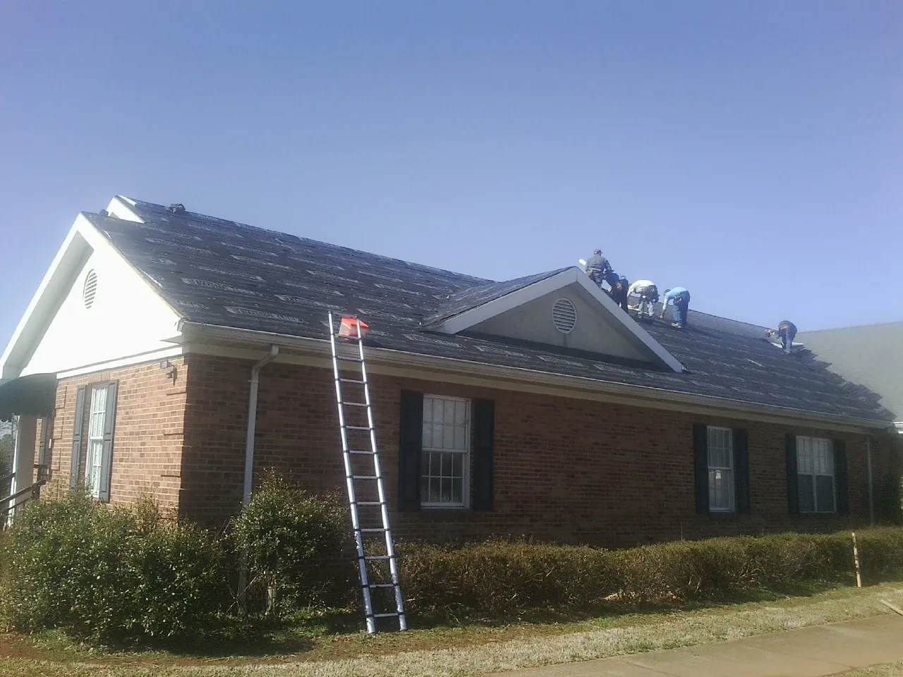 Crew tearing shingles off a brick ranch roof with a ladder set — Metal Roof Repair work by our crew in Riverview