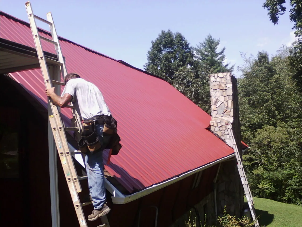 Roofer on a ladder beside a red metal roof and stone chimney — Soffit & Fascia Repair by Roofing Brothers in Riverview